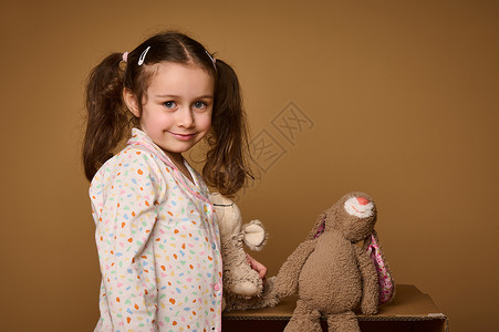 毛绒过敏特写可爱的小女孩 有两条马尾辫 拿着她的毛绒玩具 自信地看着相机 可爱地微笑着 在米色背景中被隔离 有复制广告空间背景