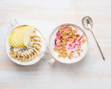 两份希腊酸奶 配果酱 苹果 麦片 奇亚籽和香蕉 放在白色木桌上的白色碗里 俯视背景图片