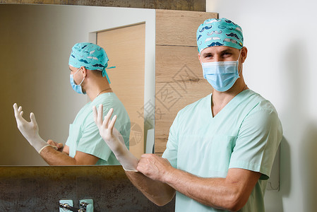 龙头面具微笑外科医生将无菌的医用手套戴在手上 并秘密地看 保护大流行的 Corona病毒为19个人提供卫生服务背景