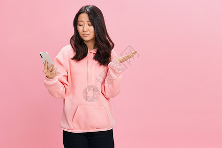 身穿粉红连帽衫的愤怒而发狂的亚洲女学生年轻女士举起手来说 在粉红工作室背景上什么姿势是孤立无助的背景图片