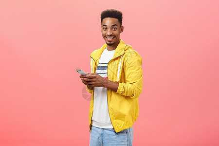 留着爆炸式卷发的时尚快乐的非洲裔美国男性半翻过粉红色的墙壁 手持智能手机 穿着黄色时髦的夹克 开心地微笑着向朋友展示设备的功能技背景图片