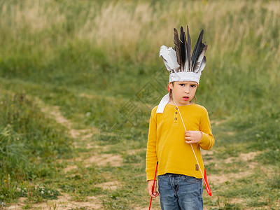拿着羽毛男孩男孩在田野上扮演美国印第安人的肖像 孩子用羽毛和弓箭手工制作头饰 演戏角色扮演 户外休闲活动背景