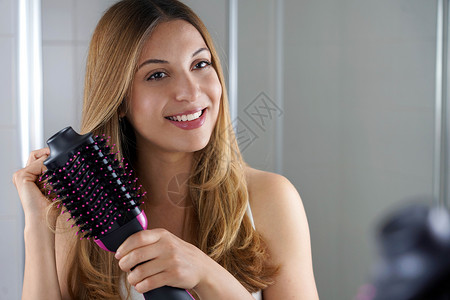 可爱的女孩拿着圆梳子烘发机 在家中的洗手间打理头发 年轻女性使用沙龙一脚吹发机和挥发器背景图片