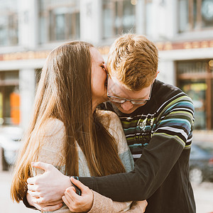 幸福的情侣在市中心拥抱的画像 戴着眼镜的红发男人亲吻着 女人留着长发 女孩在男人耳边低语 青少年爱情和初吻 爱情 关系 情侣的概背景