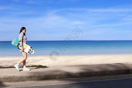 年轻女性带着冲浪滑板在热带海滩附近的小路上行走 年轻的长发女孩穿着长袖衬衫 牛仔裤和运动鞋 踩着冲浪滑板走在海滩路上青少年放松木爱好高清图片素材