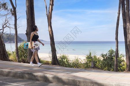 年轻女性带着冲浪滑板在热带海滩附近的小路上行走 年轻的长发女孩穿着长袖衬衫 牛仔裤和运动鞋 踩着冲浪滑板走在海滩路上女士闲暇公园旅行高清图片素材