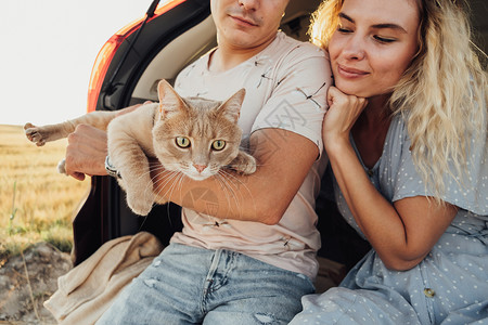 年轻女人和男人与宠物坐在汽车的Trunk 快乐夫妇与他们的红猫享受公路之旅家庭丈夫女士成人日落男朋友越野车冒险男性女性背景图片