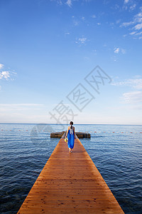 蓝色防伪码穿蓝裙子的女孩站在海边的木头码头上 有云的美丽的天空 度假概念 去欧洲旅行 (笑声)背景