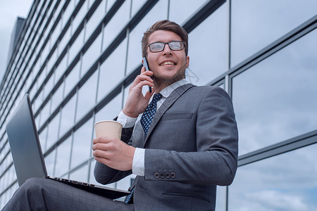坐在办公大楼旁边的青年企业公司男员工商业成人杯子手机上网咖啡男人眼镜笔记本街道背景图片