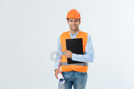 身着橙色头盔 面带微笑的年轻英俊建筑师工程师的半身肖像与蓝图合影 看着在白色背景 复制空间中隔离的相机男性项目绘画帽子建筑人士商背景图片