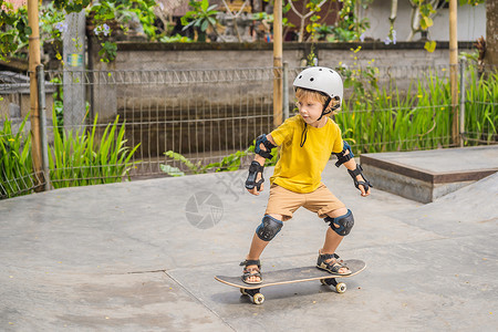 学会放下戴头盔和膝盖垫的体育男孩学会在滑板公园与滑板一起玩滑板孩子运动公园乐趣游戏男生街道平衡驾驶城市生活背景