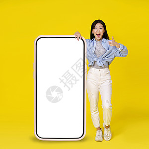 优惠大酬宾兴奋的亚洲女孩竖起大拇指 靠在巨大的 巨大的智能手机上 上面有白色 空白的屏幕 靠在黄色背景中孤立的休闲装上 自由空间模型背景