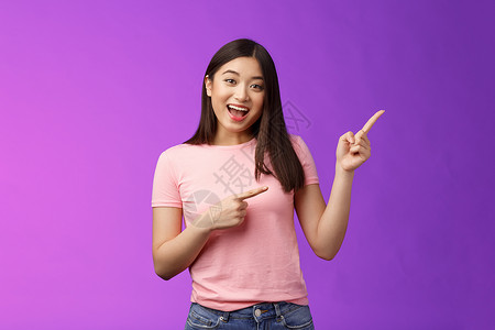 友好而热情的迷人亚洲女孩在进行有趣的谈话 讨论感兴趣的新店 向右指着邀请进来 站在紫色背景微笑的相机上旅游女朋友广告促销黑发企业背景图片