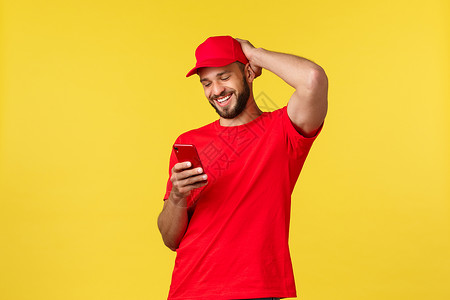 黄色载卸车非接触式支付 网上交付 网上购物和包裹跟踪概念 身穿红色制服帽和 T 恤 发短信 在黄色背景中对着智能手机微笑的快乐快递员隔离载背景