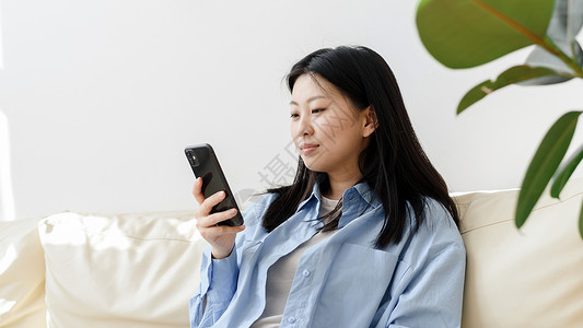 年轻的亚洲女性在客厅的沙发上放松 用智能手机浏览新闻 韩国女士在现代手机上写信息 通过网站在线购物 网页横幅背景图片