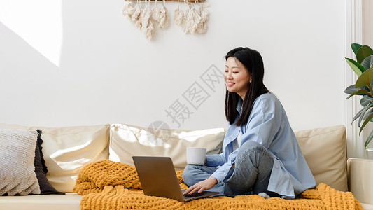 在家里舒适的客厅里 坐在沙发上使用笔记本电脑微笑的亚洲女人的画像 快乐的女学生使用电脑进行休闲 学习 教育或工作 网页横幅背景图片