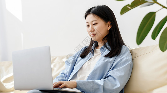 微笑的年轻亚洲女性自由职业者在家工作 坐在沙发上 使用笔记本电脑 复制空间 放松的韩国年轻女性享受周末 在笔记本电脑上上网 网页背景图片