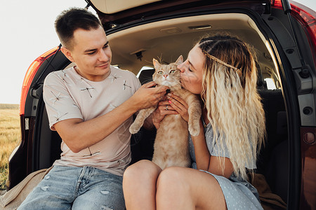 年轻女人和男人与宠物坐在汽车的Trunk 快乐夫妇与他们的红猫享受公路之旅女性女孩成人丈夫女士家庭夫妻日落妻子旅行背景图片