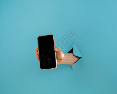 从蓝底的洞里露出的一只女性手 拿着一个带黑屏的智能手机背景图片