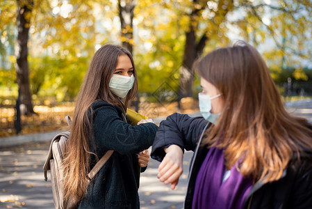 身戴保护性医疗面具的女学生朋友用手肘相互打招呼;和背景图片