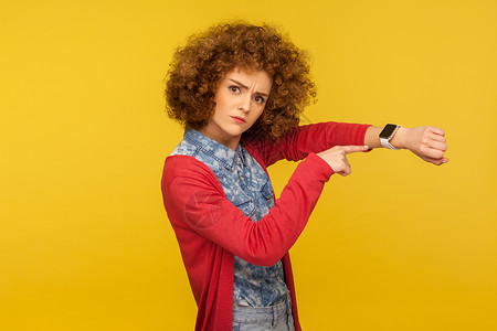 黄色背景的情绪卷发女孩的肖像老板手腕女士手指检查小时手势女性手表焦虑背景图片