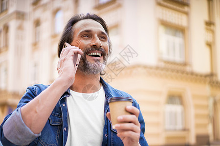 快乐旅行的成熟大胡子男人用手机说话 用一次性纸杯喝咖啡 站在户外穿着牛仔裤衬衫的旧城市背景中 自由职业者旅行人背景图片