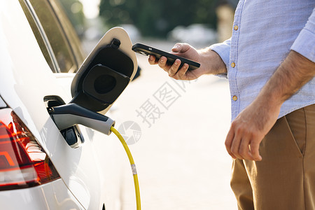 燃料汽车无法辨认的人从充电站插入电动汽车 男性使用智能手机上的应用程序将电源线插入电动汽车 商务人士在户外充电站给电动汽车充电背景