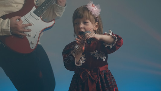 苏格兰舞穿旧礼服的小女孩在舞台上唱歌 她父亲弹电吉他嗓音青年家庭幼儿园蕾丝背景宝宝戏服爱好音乐会背景