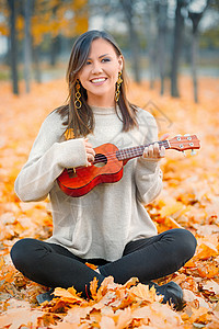 枫叶吉他素材年轻美丽的混合种族女子在秋天公园玩乌鲁莱和微笑背景