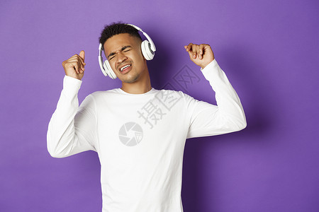 无忧无虑的非洲美国男子跳舞 用无线耳机监听音乐 站在紫底背景上促销工作室快乐广告发型工作员工手势爆炸运动衫背景图片