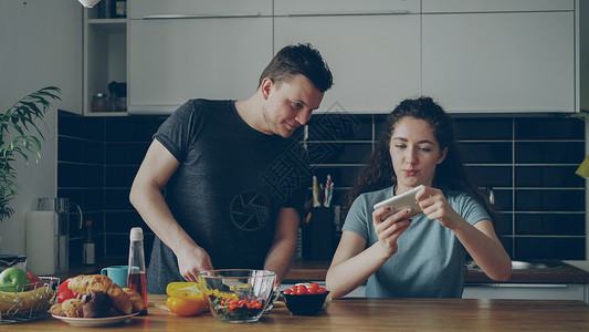 在家厨房的一对有吸引力的情侣 在她男友做饭时 卷卷发女孩在智能手机上玩电子游戏背景图片