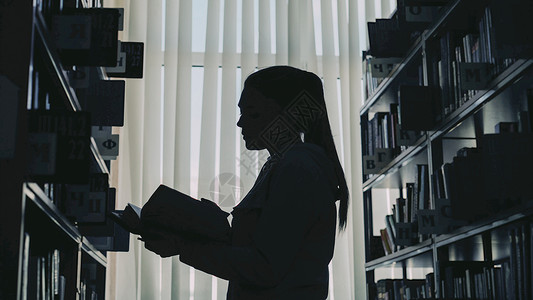 年轻女学生的月光在大型图书馆书架上站立 反对持有书本翻页和阅读的窗窗壁背景图片