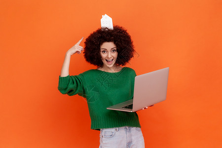 编程爆炸素材带着非洲发型的有趣兴奋女人穿着绿色散装毛衣 用笔记本电脑指着纸屋头顶上背景