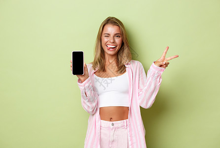 吃惊女穿着粉红色衬衫的金发美女肖像 展示和平标志和手机屏幕 微笑喜悦 站在绿色背景上站立背景