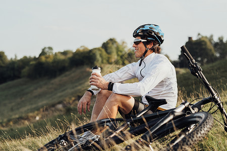 瓶子里的水职业男性骑自行车的人从瓶子里喝水 男人在乡下户外旅行时坐在自行车旁车轮运动员踪迹山地运动闲暇冒险眼镜速度太阳镜背景