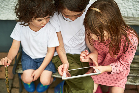 三名可爱的小孩在户外坐在长椅上时使用数码平板电脑 他们用数字平板电脑做客背景图片