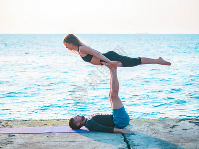 一起去海边在海边做Acroyoga的年轻夫妇 男人躺在混凝土板上 让女人靠在脚上平衡 漂亮的一对一起练瑜伽练习朋友们花园女性冒充力量运动身背景