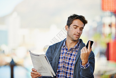 一个英俊的男人拿着报纸 在户外使用他的手机 校对背景图片