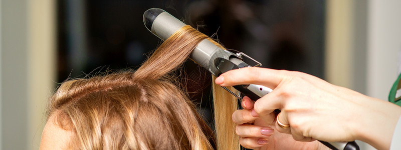 理发工具带发型师在美容院为长棕色头发的年轻女子做卷发卷发和卷心铁客厅金子理发师梳妆台沙龙工作黑发女孩客户工具背景