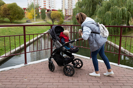 母亲婴儿车一名年轻母亲肩上背着背包 带着孩子在市公园的婴儿园里散步背景