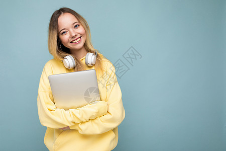 黄色耳机身穿黄色连帽衫的欧洲年轻快乐微笑的女学生手持笔记本 背景为孤立的蓝色背景 有可用空间 教育理念电脑情怀耳机女性成人学生自由职业者背景