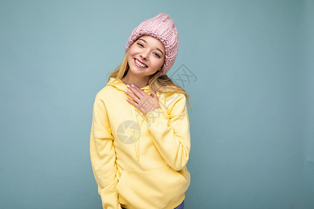 黄色帽衫迷人的微笑快乐的金发年轻女性孤立地站在色彩缤纷的背景墙上 穿着日常时尚的服装 看着镜头时表现出面部情绪金发女郎帽子黄色粉红色帽衫背景
