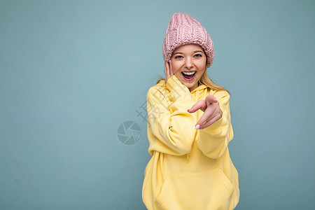 粉色薄衫照片中 美丽 微笑的年轻金发女子被隔离在蓝色背景墙上 戴着冬季粉色帽子和黄色连帽衫 看着相机 指着你背景