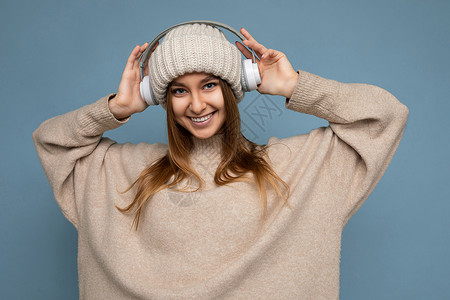 特写照片美丽迷人积极快乐微笑的年轻深色金发女人穿着米色时尚毛衣和针织米色冬季帽子隔离在蓝色背景与复制空间戴着白色蓝牙无线耳机 音背景图片
