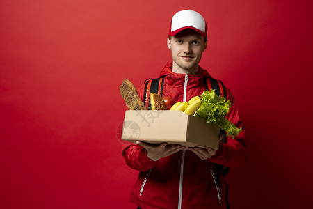 开朗英俊的年轻送货员拿着杂货店购物盒的画像 背景是红色的 网上购物 餐厅的优质服务 现代购物理念背景图片
