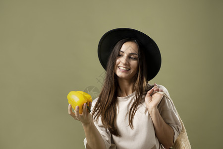 戴着黑色帽子的黑发女人微笑着 手里拿着购物网生态袋 上面装着有机蔬菜 手里拿着黄胡椒 享受健康生活购物背景图片