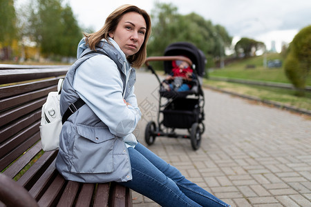 远离抑郁素材恼怒的年轻母亲坐在公园的长椅上 远离婴儿车 脸上带着愤怒的表情 产后抑郁症的概念背景