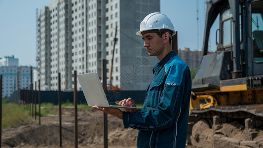 我是一名工程师一名戴头盔的男性建筑师站在一个建筑工地的背景之下 手里拿着一台笔记本电脑基础设施检查领班脚手架蓝图安全帽工程师承包商房子企业家背景