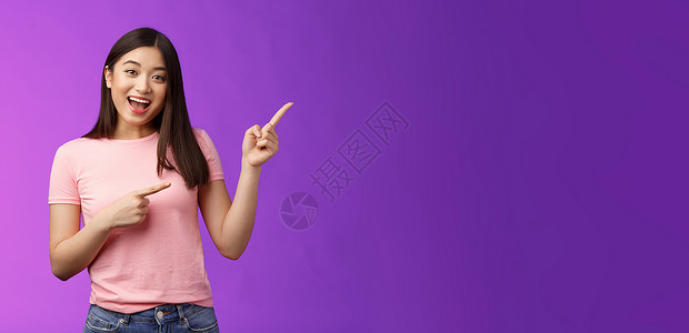 友好而热情的迷人亚洲女孩在进行有趣的谈话 讨论感兴趣的新店 向右指着邀请进来 站在紫色背景微笑的相机上展示黑发情感促销大学旅游学背景图片