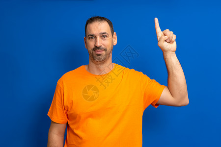 40 多岁有见地 聪明 积极主动的白人男子 身穿橙色 T 恤 举着食指 提出了与蓝色背景工作室肖像隔离的伟大新想法 人们的生活方背景图片